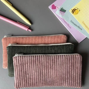May include: Three corduroy pencil cases in shades of pink and green, with a zip closure. Two pens and a notepad with a bee illustration are also visible. The pencil cases are rectangular and have a ribbed texture.