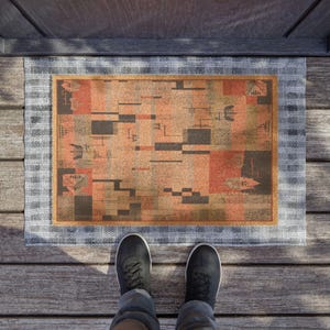 May include: A rectangular doormat featuring a geometric pattern in orange, brown, and black. It has a light blue and white checkered border and a gold-coloured inner border. The image is a top-down view, showing the mat on a wooden surface.