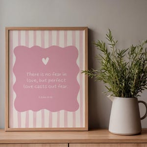 Puede incluir: Un estampado enmarcado con un fondo a rayas rosas y blancas. El estampado presenta un diseño rosa en forma de nube con un corazón blanco y el texto "No hay temor en el amor, sino que el amor perfecto echa fuera el temor." Una jarra blanca con vegetación está a la derecha.