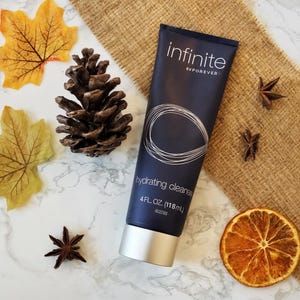 May include: A navy blue tube of "infinite" hydrating cleanser, 4 fl oz (118 ml), with a silver cap. The tube features a white circular design. The image includes fall foliage, a pine cone, star anise, and a dried orange slice.