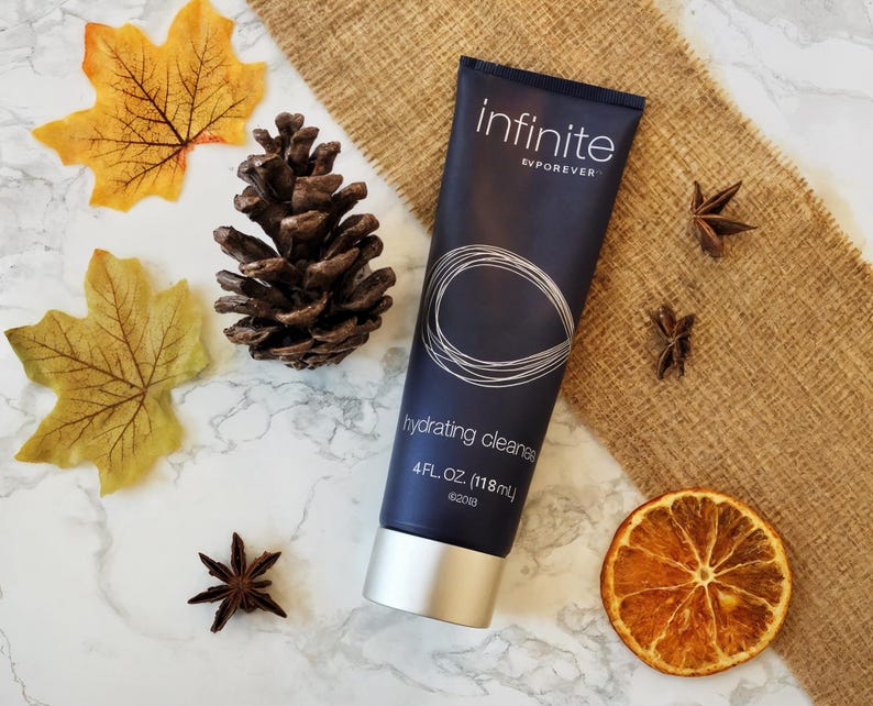 May include: A navy blue tube of "infinite" hydrating cleanser, 4 fl oz (118 ml), with a silver cap. The tube features a white circular design. The image includes fall foliage, a pine cone, star anise, and a dried orange slice.