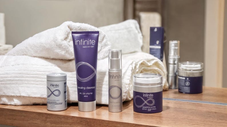 May include: A collection of skincare products, including a cleanser, serum, and cream, arranged on a wooden surface. The products are in purple and silver packaging, with the brand name "infinite" and a stylized infinity symbol. White towels are in the background.