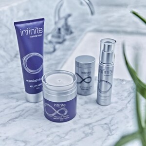 May include: A collection of skincare products, including a tube, a jar, and two cylindrical containers, arranged on a marble surface. The products are primarily purple and silver, with the word "infinite" and a stylized infinity symbol prominently displayed. Green leaves are in the background.