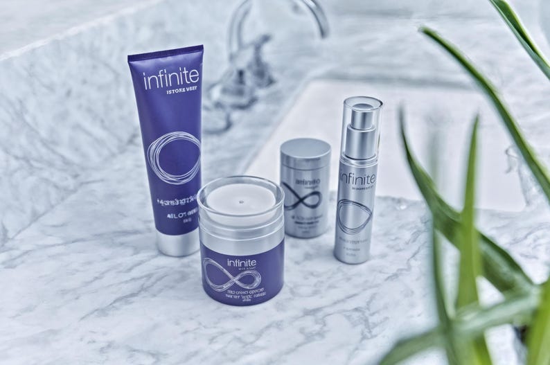 May include: A collection of skincare products, including a tube, a jar, and two cylindrical containers, arranged on a marble surface. The products are primarily purple and silver, with the word "infinite" and a stylized infinity symbol prominently displayed. Green leaves are in the background.