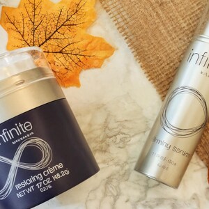 May include: Two skincare products, a jar of restoring cr&egrave;me and a bottle of firming serum, are displayed with fall foliage. The jar is navy blue with the word "infinite" and the serum is silver. A dried orange slice is also visible.