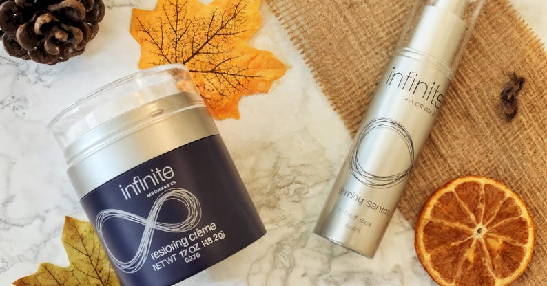 May include: Two skincare products, a jar of restoring cr&egrave;me and a bottle of firming serum, are displayed with fall foliage. The jar is navy blue with the word "infinite" and the serum is silver. A dried orange slice is also visible.
