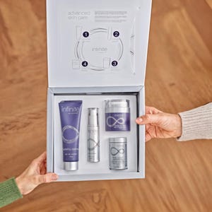 May include: A white box set of skincare products, held by two hands. The box is open, revealing four products: a purple tube, a silver pump bottle, a purple jar, and a silver container. The products have the brand name "infinite" and a swirling design.