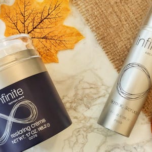 May include: Two skincare products, a jar and a bottle, are displayed on a marble surface with autumn leaves and a dried orange slice. The jar is dark blue with the word "infinite" and "restoring cremes". The bottle is silver with the word "infinite" and "firming serum".