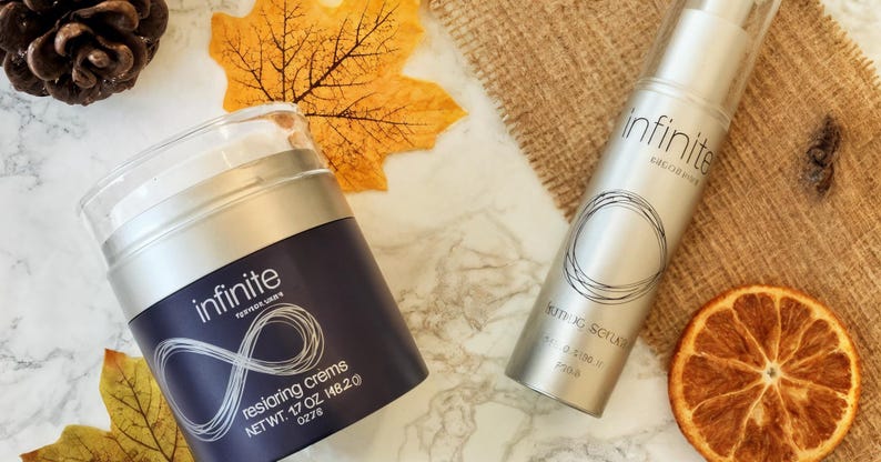 May include: Two skincare products, a jar and a bottle, are displayed on a marble surface with autumn leaves and a dried orange slice. The jar is dark blue with the word "infinite" and "restoring cremes". The bottle is silver with the word "infinite" and "firming serum".