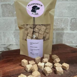 May include: A package of PupPower Pure salmon cubes dog treats. The treats are freeze-dried and appear as small, beige cubes. The package has a circular label with a black dog silhouette and the words "PupPower Pure".