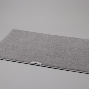 May include: A rectangular grey bath mat with a textured, looped pile surface and a finished border. A small white fabric label with black text is sewn onto the bottom edge of the mat, featuring the brand name Shuttle Fine in a clear font.