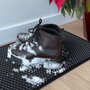 May include: Brown leather hiking boots, dusted with snow, sit on a black textured mat. The boots feature black laces and a robust sole. A red poinsettia plant is partially visible in the background, suggesting a winter scene.
