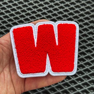 May include: A red, embroidered letter "W" patch with a white border. The letter has a textured, fuzzy appearance. The patch is likely for clothing or accessories.