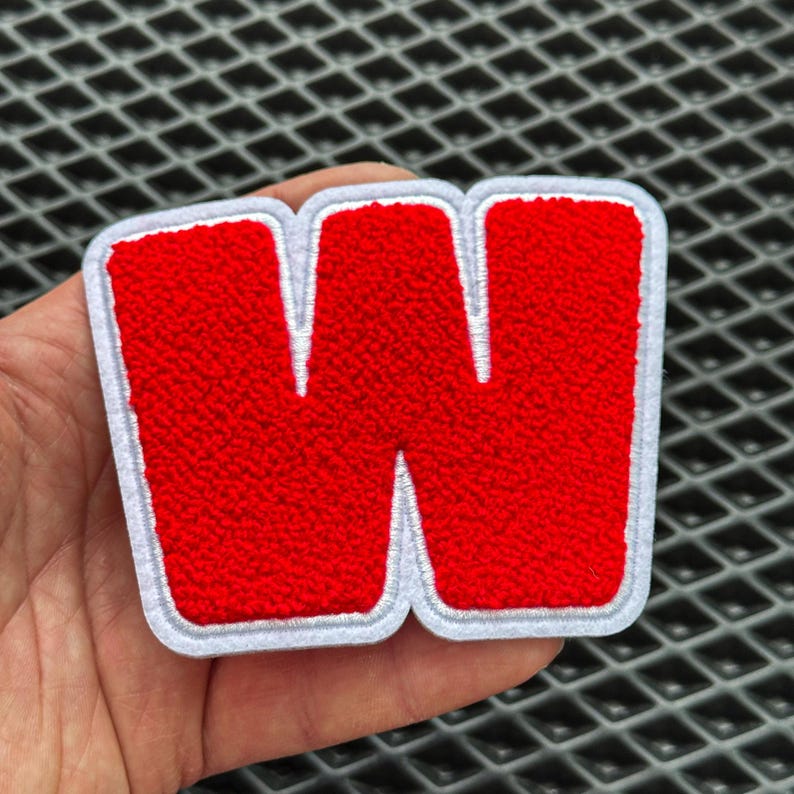May include: A red, embroidered letter "W" patch with a white border. The letter has a textured, fuzzy appearance. The patch is likely for clothing or accessories.