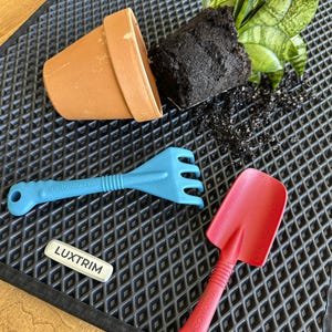 May include: A terracotta flower pot, blue rake, and red trowel are arranged on a textured surface. A small plant with green leaves and soil is next to the pot. A white label with the text "LUXTRIM" is also present.