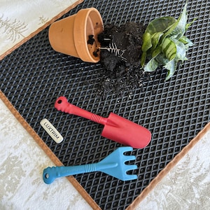 May include: A terracotta pot, soil, and a green plant are on a black textured mat. A red trowel and a blue hand rake are also present. A white label with the text "LUXTRIM" is visible.