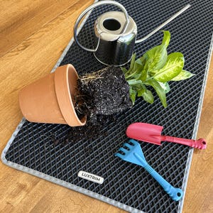 May include: A terracotta pot with soil and a plant overturned on a black mat. A silver watering can, a pink trowel, and a blue rake are also present. The mat has a diamond pattern and the brand name "LUXTRIM" is visible.