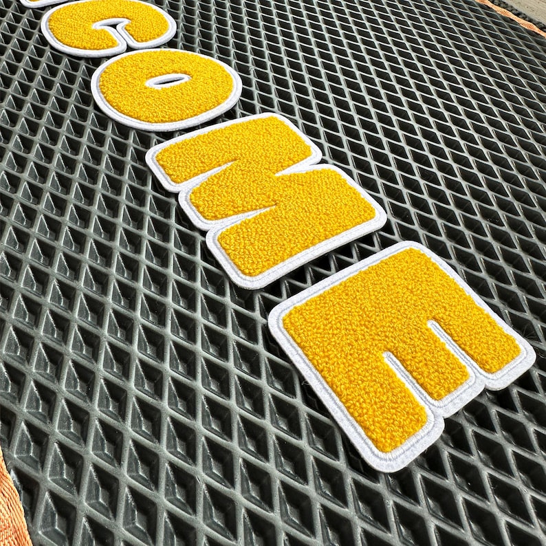 May include: Yellow chenille patches spelling "COME" with a white border. The patches are on a grey diamond-patterned surface. The letters are in a bold, block style.