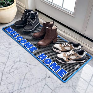 May include: A rectangular doormat with the words "WELCOME HOME" in blue lettering. The mat is black with a blue border. Various pairs of shoes and boots are placed on the mat, including black boots, brown boots, and trainers.
