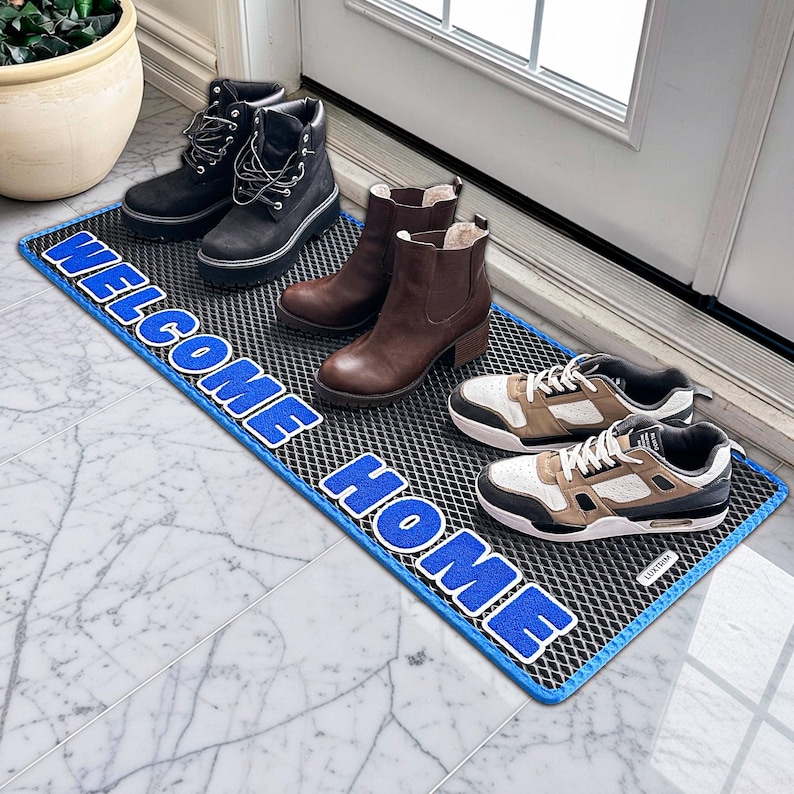 May include: A rectangular doormat with the words "WELCOME HOME" in blue lettering. The mat is black with a blue border. Various pairs of shoes and boots are placed on the mat, including black boots, brown boots, and trainers.