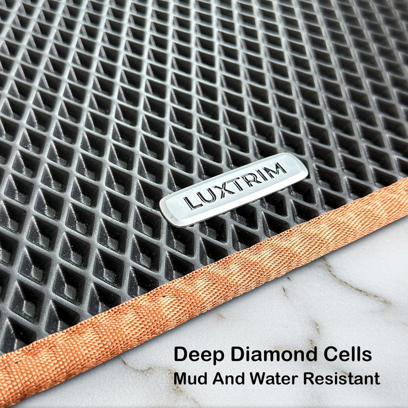 May include: A close-up of a grey car floor mat with a diamond pattern. The mat features a silver "LUXTRIM" emblem and a copper-coloured border. Text at the bottom reads "Deep Diamond Cells Mud And Water Resistant."