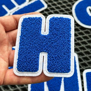 May include: A blue letter "H" patch with a white border. The patch has a textured, felt-like appearance. Other letter patches are visible in the background. This patch is suitable for jackets, bags, or other fabric items.