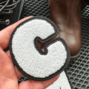 May include: A white, textured letter "C" patch with a black border. The patch is held in front of a pair of black shoes and a brown boot. The patch is likely for a jacket or bag.