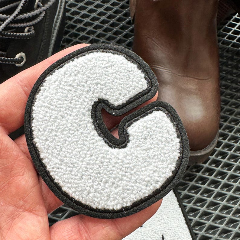 May include: A white, textured letter "C" patch with a black border. The patch is held in front of a pair of black shoes and a brown boot. The patch is likely for a jacket or bag.