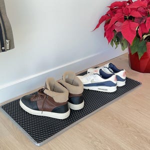 May include: A black and grey textured doormat with a silver border holds three pairs of shoes. The shoes include brown and black high-top trainers, and white and blue trainers. A red poinsettia plant is in the background.
