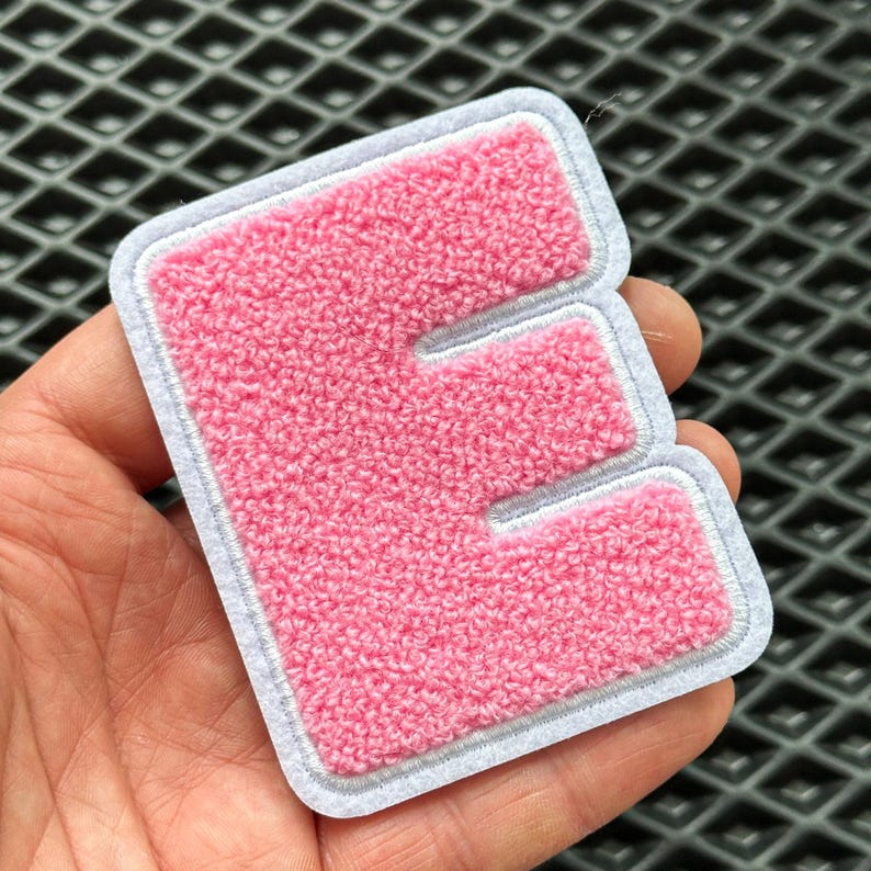 May include: Pink chenille letter "E" patch with a white border and grey stitching. The letter has a textured, fuzzy appearance. This embroidered patch can be used to personalise clothing, bags, or other fabric items.