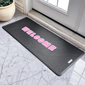 May include: A black rectangular doormat with a diamond pattern. The word "WELCOME" is printed in large, pink, block letters across the centre. The mat is placed in front of a white door on a marble tile floor.