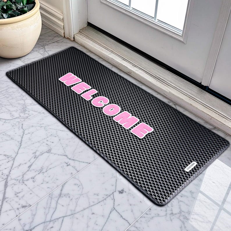 May include: A black rectangular doormat with a diamond pattern. The word "WELCOME" is printed in large, pink, block letters across the centre. The mat is placed in front of a white door on a marble tile floor.