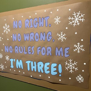 May include: A brown poster with the text "NO RIGHT, NO WRONG, NO RULES FOR ME I'M THREE!" in blue and purple, with white snowflake and dot decorations. The poster is mounted on a green wall.