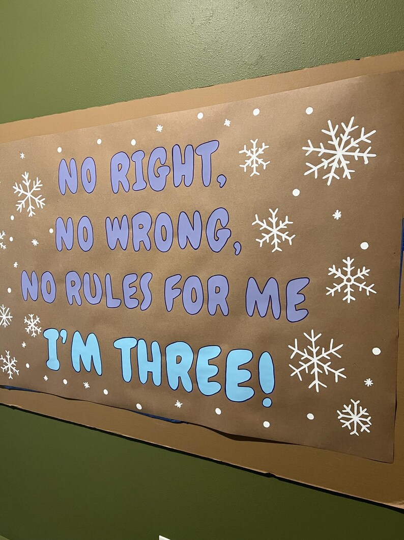 May include: A brown poster with the text "NO RIGHT, NO WRONG, NO RULES FOR ME I'M THREE!" in blue and purple, with white snowflake and dot decorations. The poster is mounted on a green wall.