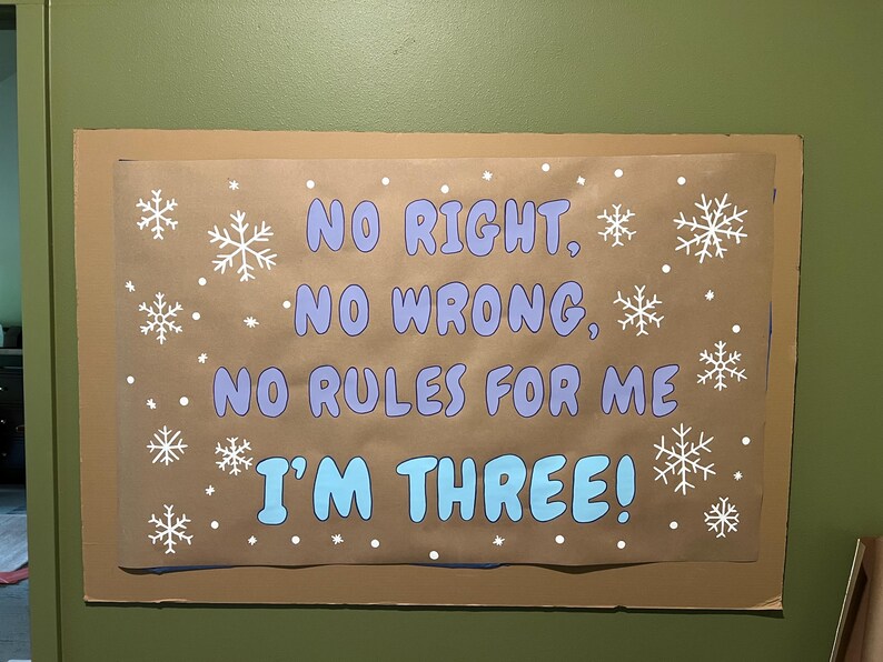 May include: A brown paper sign with the text "NO RIGHT, NO WRONG, NO RULES FOR ME I'M THREE!" in blue and white. White snowflakes and dots are scattered across the sign.