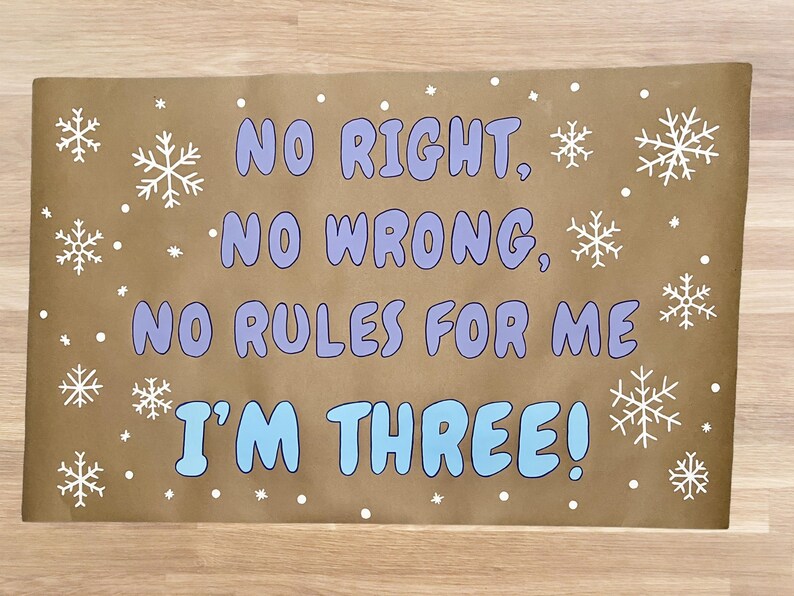 Puede incluir: Un letrero rectangular marr&oacute;n con el texto "NO RIGHT, NO WRONG, NO RULES FOR ME I'M THREE!" en azul y morado. Copos de nieve y puntos blancos decoran el letrero.