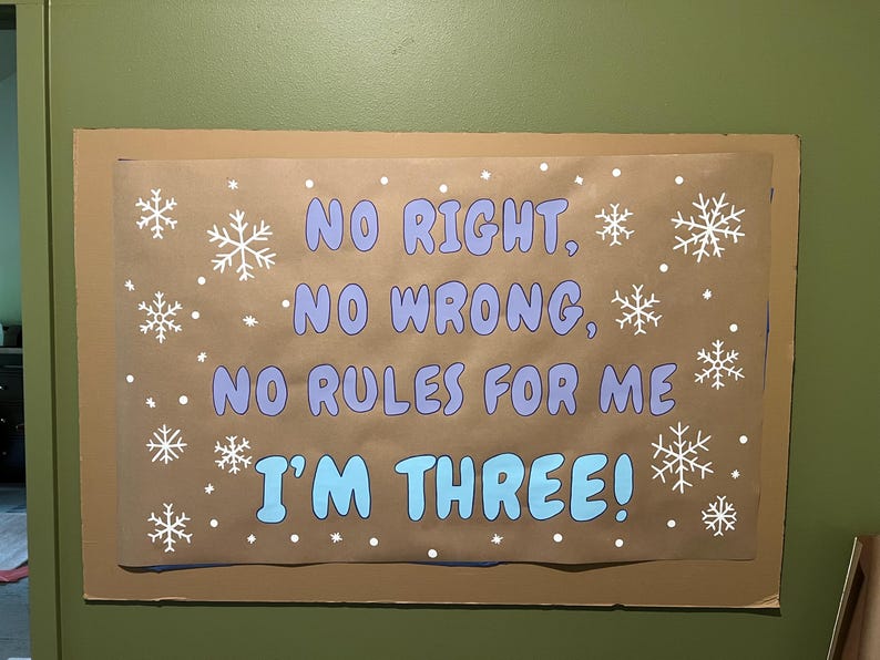Puede incluir: Un letrero de papel marr&oacute;n con el texto "NO RIGHT, NO WRONG, NO RULES FOR ME I'M THREE!" en azul y blanco. Copos de nieve y puntos blancos est&aacute;n dispersos por el letrero.