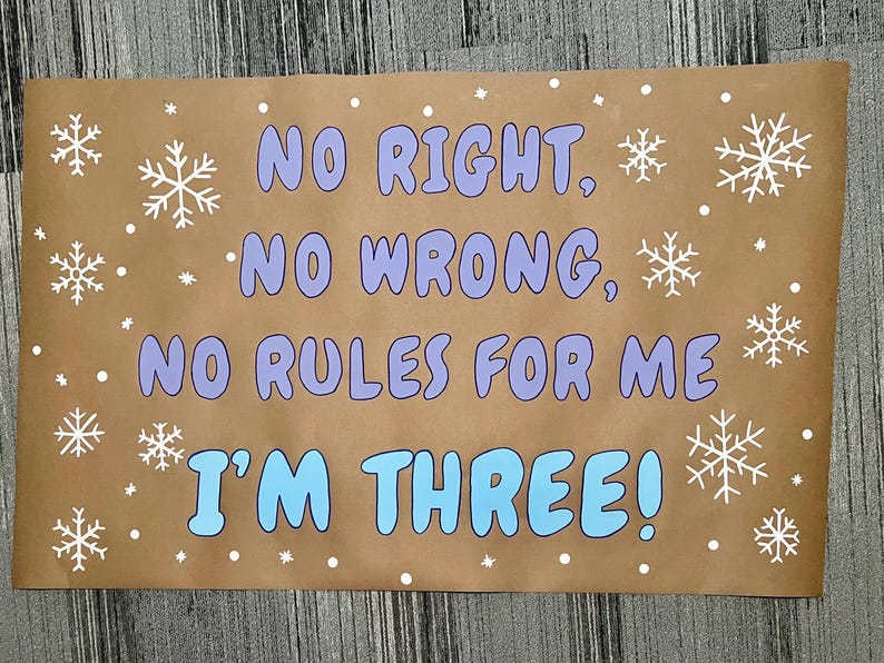Puede incluir: Un letrero de papel marr&oacute;n con el texto "NO RIGHT, NO WRONG, NO RULES FOR ME I'M THREE!" en letras azules y moradas. Copos de nieve y puntos blancos rodean el texto.
