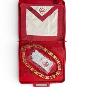 May include: A red case containing Masonic regalia. Inside, a white apron with a red border and emblem, a gold-colored chain, white gloves, and a red emblem are visible. The case is open, revealing the contents.