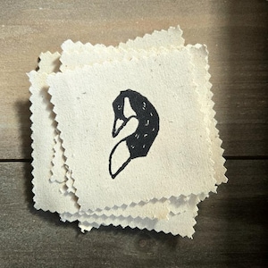 May include: A stack of small, square, off-white fabric patches with a black goose head design. The patches have a raw, unfinished edge and are stacked on a wooden surface.