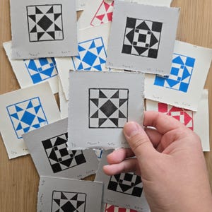 May include: A selection of small, square art prints with geometric designs in black, blue, and red ink on textured paper. The prints display star and diamond patterns. One print is held by a hand.