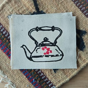 May include: A rectangular piece of off-white canvas fabric featuring a black line drawing of a tea kettle. The kettle design includes a handle, a spout, a lid, and a small cluster of five red floral shapes printed on the center of the body.