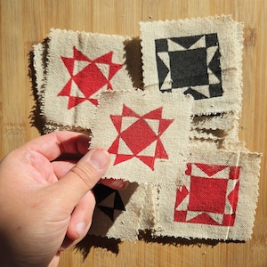 May include: A collection of square, raw-edged beige fabric patches featuring printed geometric star patterns in solid red and black ink. The patches are stacked, with one red star patch held up to display the textured weave of the natural fabric.
