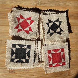May include: Four square burlap coasters with a rustic, frayed edge. Each coaster features a geometric star or square design in red or black. The coasters are arranged on a wooden surface.