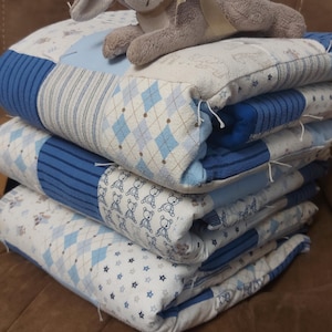 May include: Four baby quilts in blue and white, patterned with diamonds, stars, and teddy bears. A grey plush bunny with a cream bow sits atop the quilts. The quilts are stacked on top of each other.