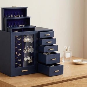 May include: A navy blue jewelry organizer with multiple compartments and drawers. The organizer has a glass-fronted section for hanging jewelry and several small drawers. It sits on a wooden surface, with a perfume bottle and a small dish nearby.