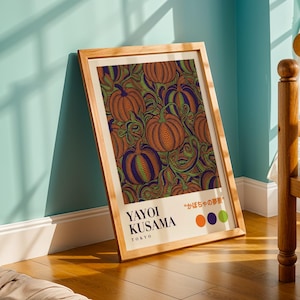 May include: A framed art print with a repeating pattern of orange pumpkins with green and purple details. The print includes the text "YAYOI KUSAMA TOKYO" and Japanese text. The frame is made of light-coloured wood.