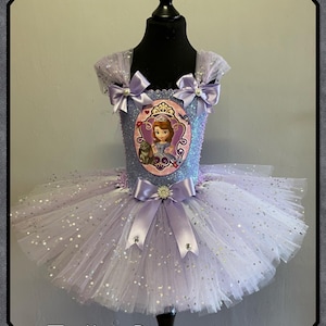 May include: A lavender and white tutu dress with a bodice featuring a princess graphic. The dress has a layered tulle skirt with silver glitter accents. Lavender bows adorn the shoulders and waist. The text "Tulle Boutique" is at the bottom.