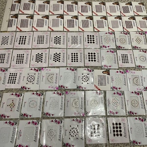 May include: A selection of packaged bindi dots in various designs and colours, including black, red, and clear rhinestones. The packages are labelled with the brand names "Aishwarya" and "Lazree".