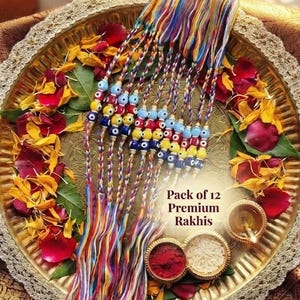 May include: A pack of 12 premium Rakhis featuring braided multi-colored threads and blue, yellow, and white evil eye beads. The text Pack of 12 Premium Rakhis is visible on the image. The items are arranged on a round gold-toned metal tray.